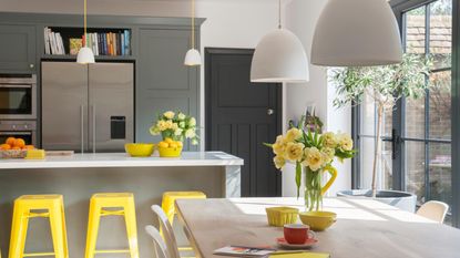 Contemporary kitchen diner. Charcoal grey units and island unit, vibrant yellow bar stools, wood dining table, white pendants, bifold doors.
