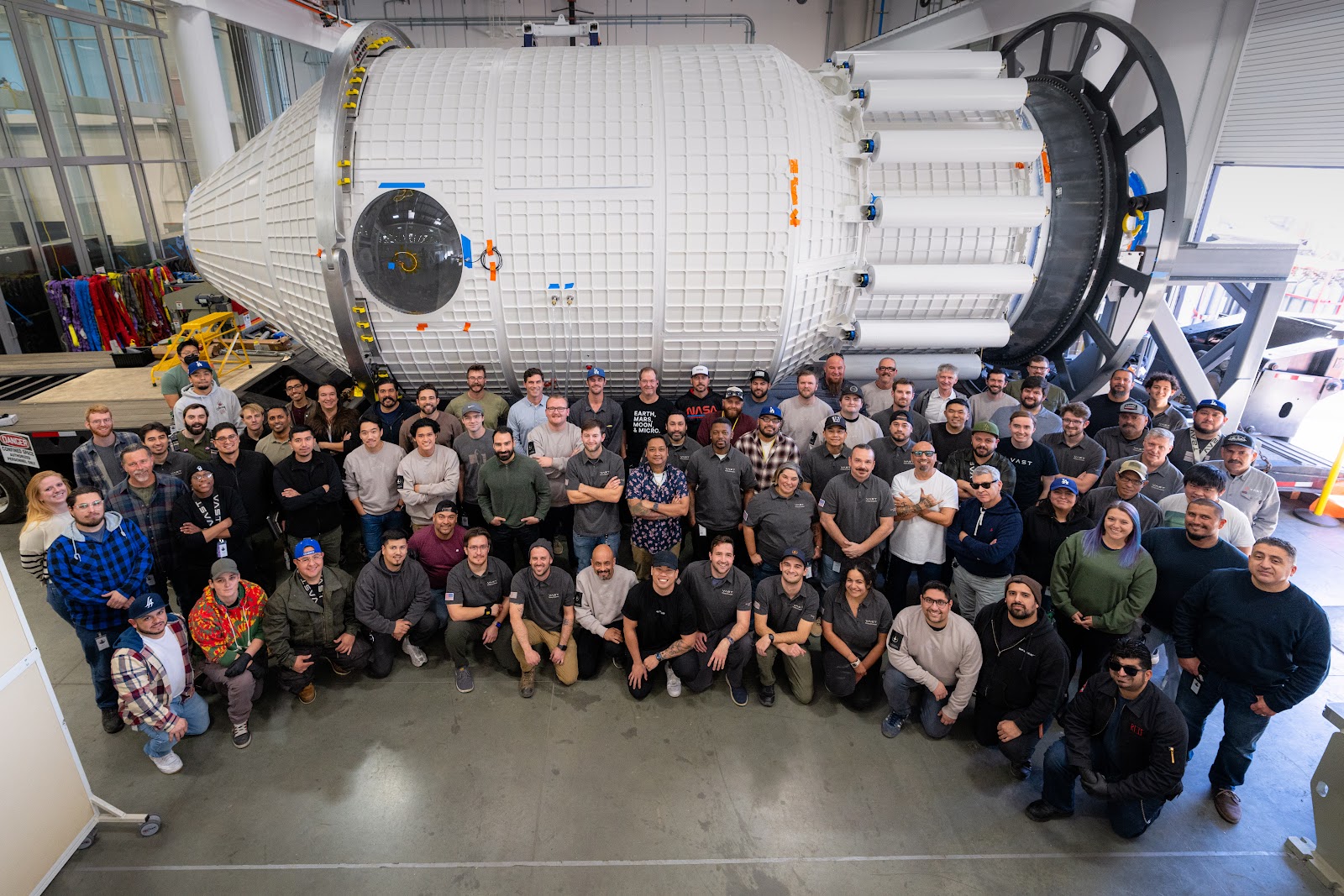 A large group of people kneel and stand in three-ish rows, in front of a large white space station module, laid on its side in a large factory.