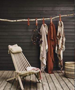 A garden chair with cushions, and three wool blankets hung from a branch on a deck outside a building.