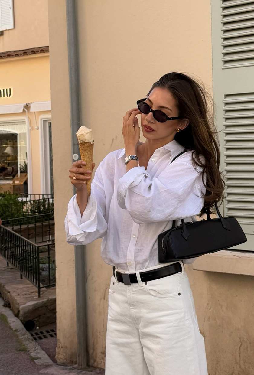 A woman standing outside with ice cream, wearing a white button-down shirt, black belt, white jeans, black sunglasses, silver jewelry, and a black shoulder bag