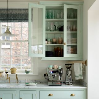 Mint green painted kitchen with glazed wall unit and coffee station set up on worktop