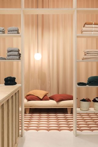 A warmly lit shop with retro-inspired, pink and wood-shaded decor shows folded jumpers on minimal shelving units and a beige futon covered in orange and yellow pillows.