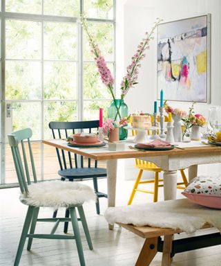 Kitchen table on a white painted wooden floor with painted wooden chairs and vases of pink and yellow flowers.
