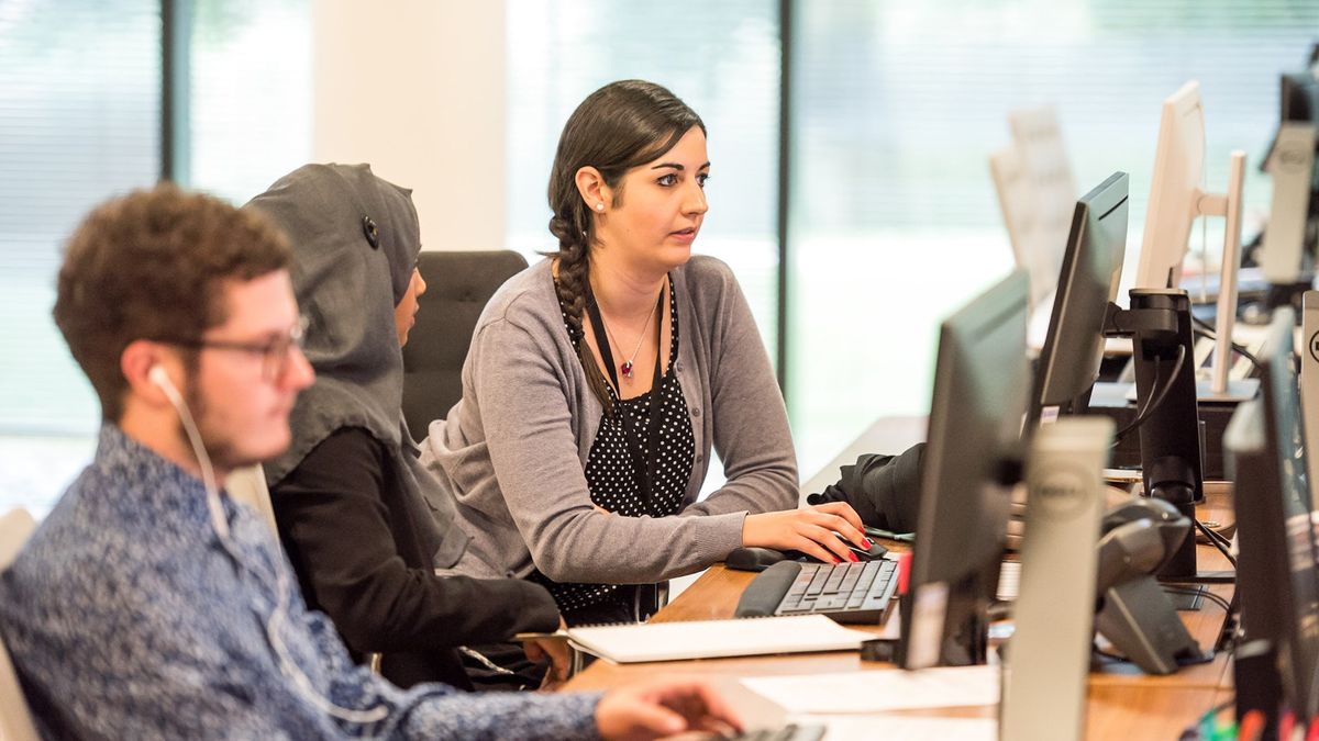 woman seated at a shared work table at an office showing her coworker something on a desktop monitor