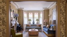 Elegant living room framed by doorway, featuring blue sofas, a bay window with stained glass details, an upholstered ottoman, a green armchair, and an upright piano by the fireplace.