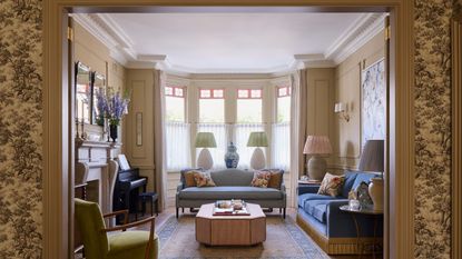 Elegant living room framed by doorway, featuring blue sofas, a bay window with stained glass details, an upholstered ottoman, a green armchair, and an upright piano by the fireplace.