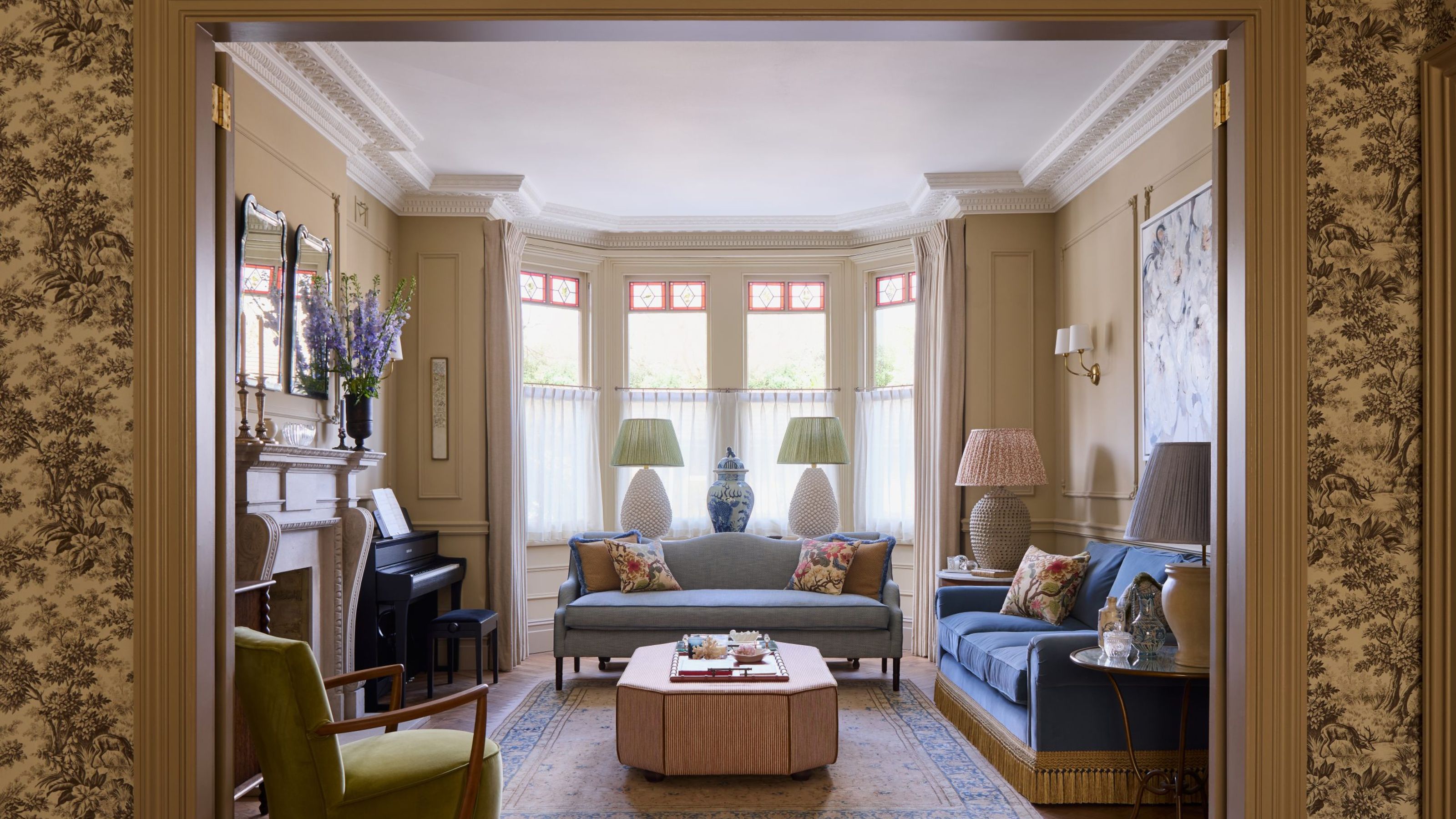 Elegant living room framed by doorway, featuring blue sofas, a bay window with stained glass details, an upholstered ottoman, a green armchair, and an upright piano by the fireplace.