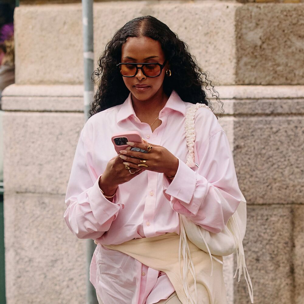 A woman in a pink button-down, white shoulder bag, sweater tied around the waist, and sunglasses, looking at their phone