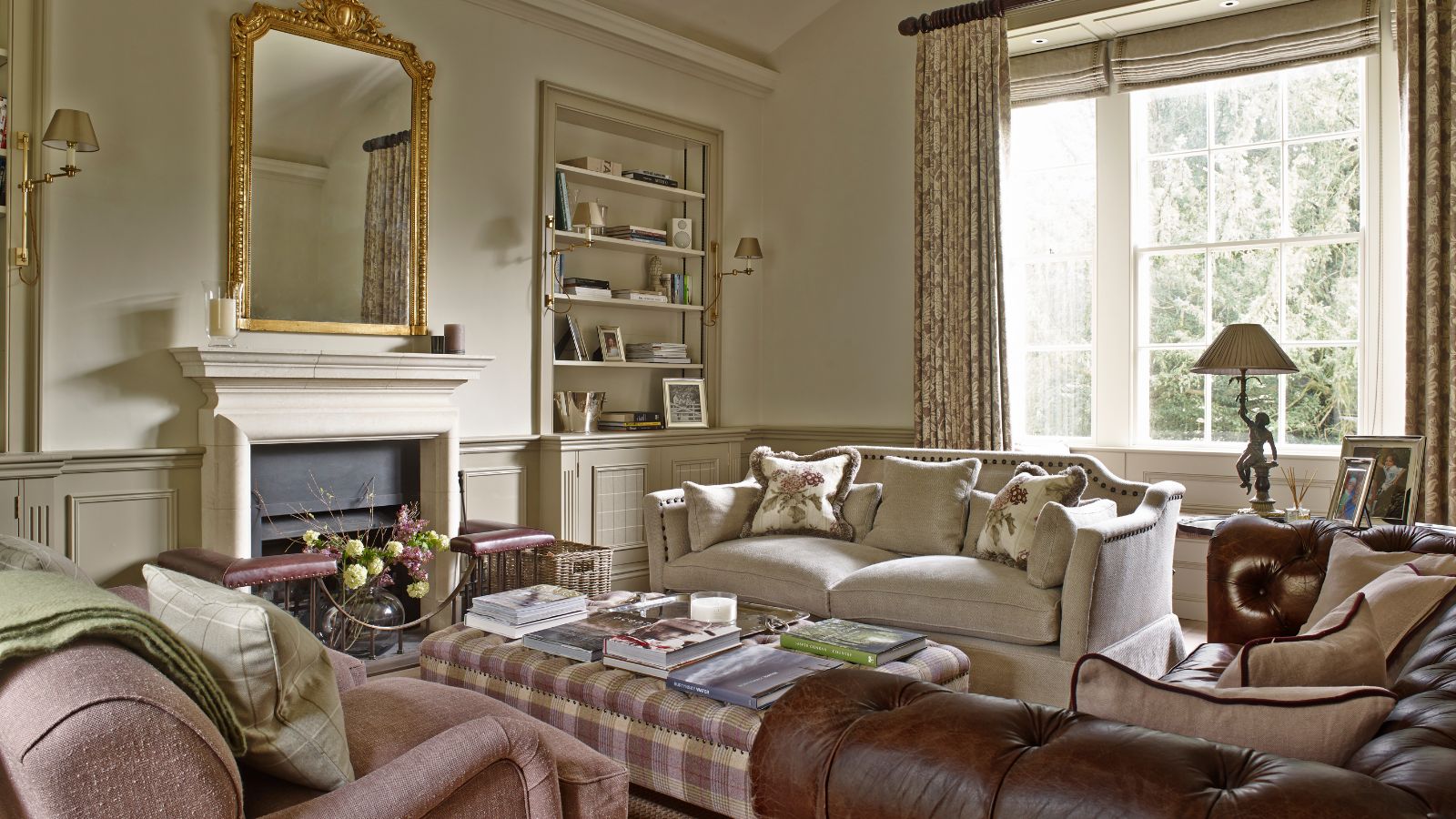 A traditional style living room with white walls, a gold mirror, patterned, mismatched sofas, a plaid ottoman, large windows, and open curtains.
