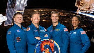 four smiling people — three men and one woman — in blue flight suits stand in front of an image of earth at night, captured from low earth orbit.