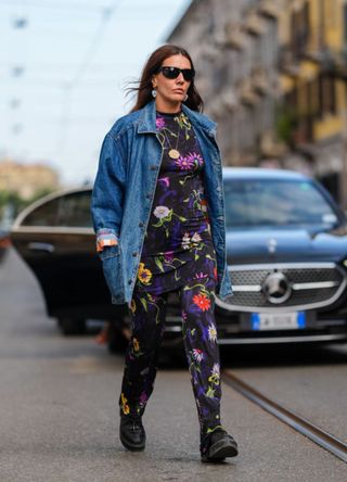 A guest wears black sunglasses, silver earrings, black floral graphic pattern/print mini dress, black floral graphic pattern/print pants, light blue oversized denim jean jacket, black platform leather boots, outside Missoni, during the Milan Fashion Week Spring/Summer 2024-2025 on September 20, 2024 in Milan, Italy.