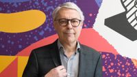 Claude Guillemot, wearing a suit and glasses, stands in front of a brightly coloured patterned background.
