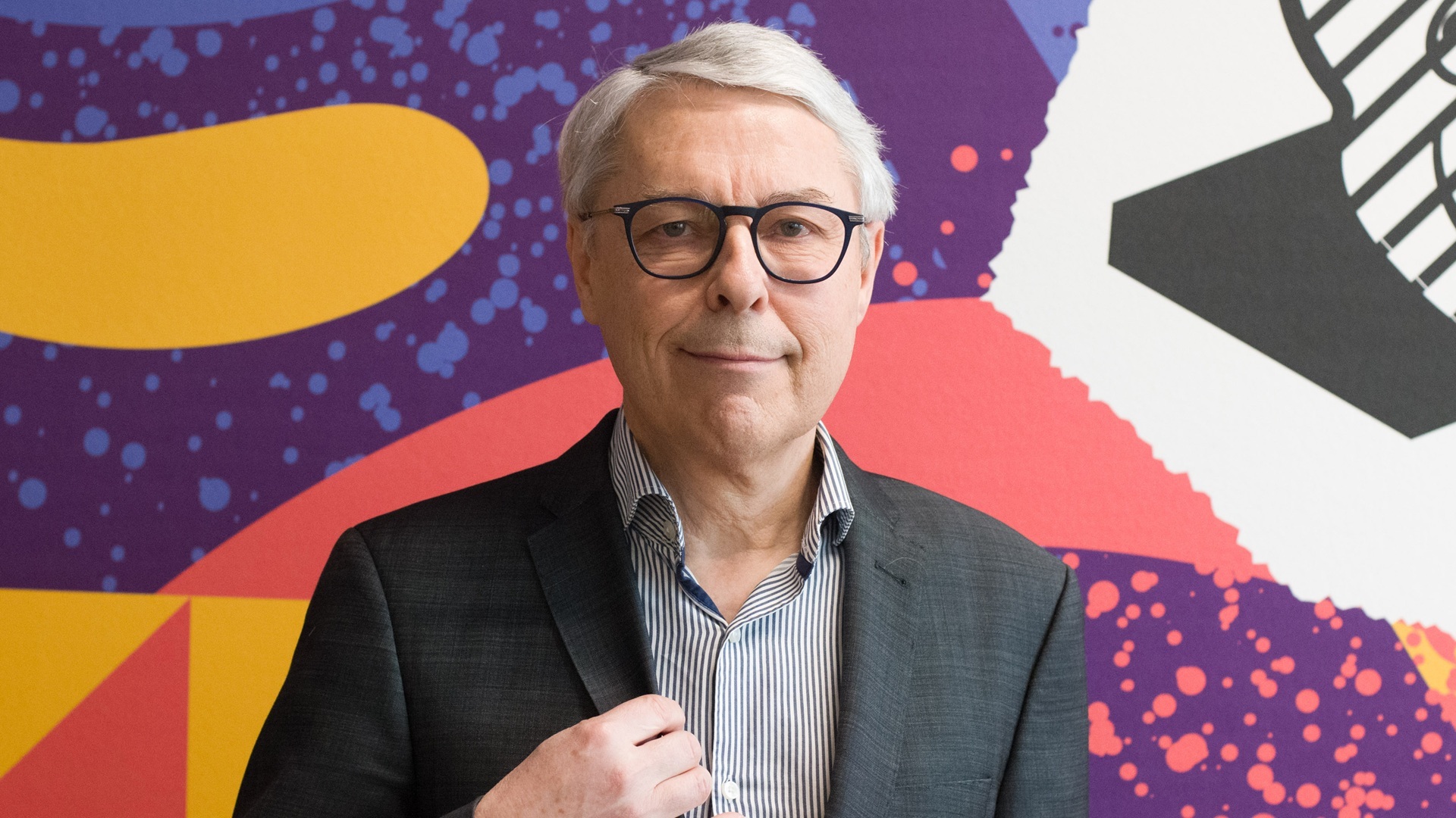 Claude Guillemot, wearing a suit and glasses, stands in front of a brightly coloured patterned background.