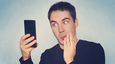 Closeup portrait of handsome young man shocked surprised, open mouth and eyes, by what he sees on his cell phone, on blue background. Negative human emotion facial expression feeling