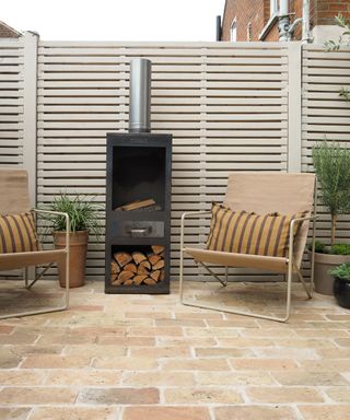 A cream painted fence with neutral brick flooring and a cream chair with log burning outdoor stove