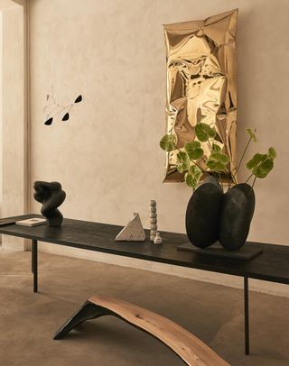 Image of a beige and stone entryway with a piece of golden art on the wall that resembles a rectangular deflated balloon. There is a long, black console table with two organically-shaped vases on it and three white decor objects in the center.