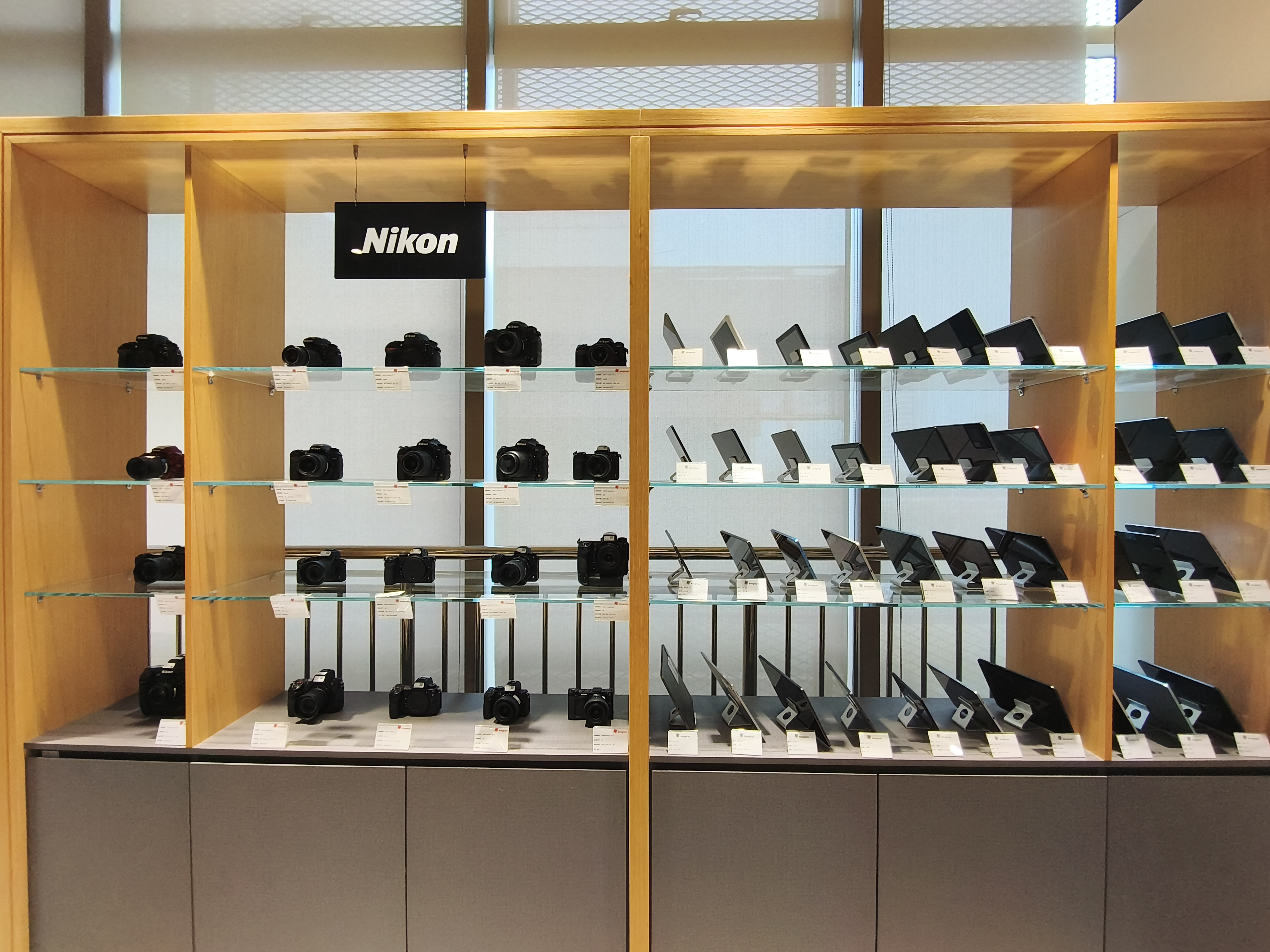 A wooden retail display case features several rows of Nikon DSLR cameras on the left and various tablet devices on the right.