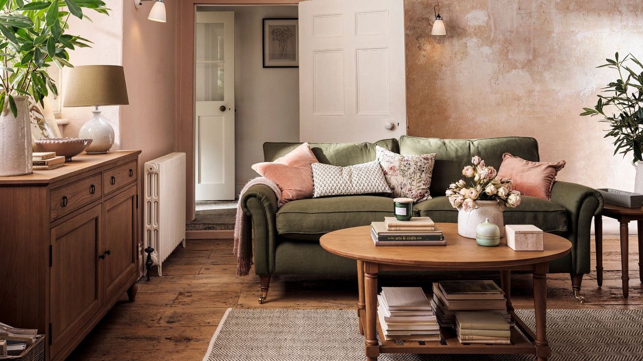 A green velvet sofa with pink floral cushions on it. A round wooden coffee table in front with books stacked beneath it. 