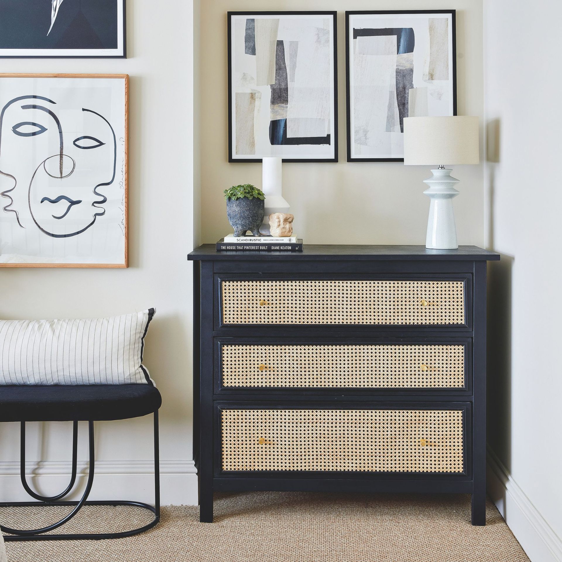 Biege painted bedroom with a black chest of drawers with rattan frontage, with decor on top it and artwork on the wall