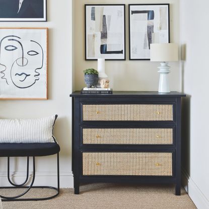 Biege painted bedroom with a black chest of drawers with rattan frontage, with decor on top it and artwork on the wall