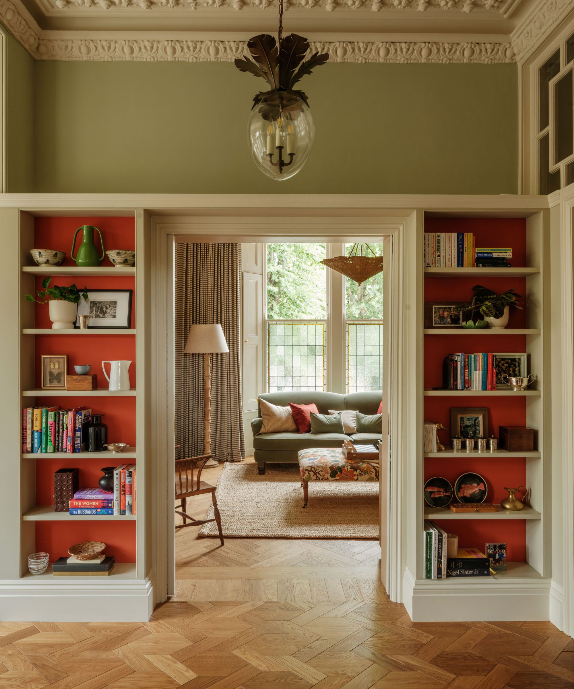 Living room with built-in bookshelves painted bright red, looking into a larger living room with a sofa, parquet flooring, large windows with shutters, a floral ottoman, and a wooden floor lamp.
