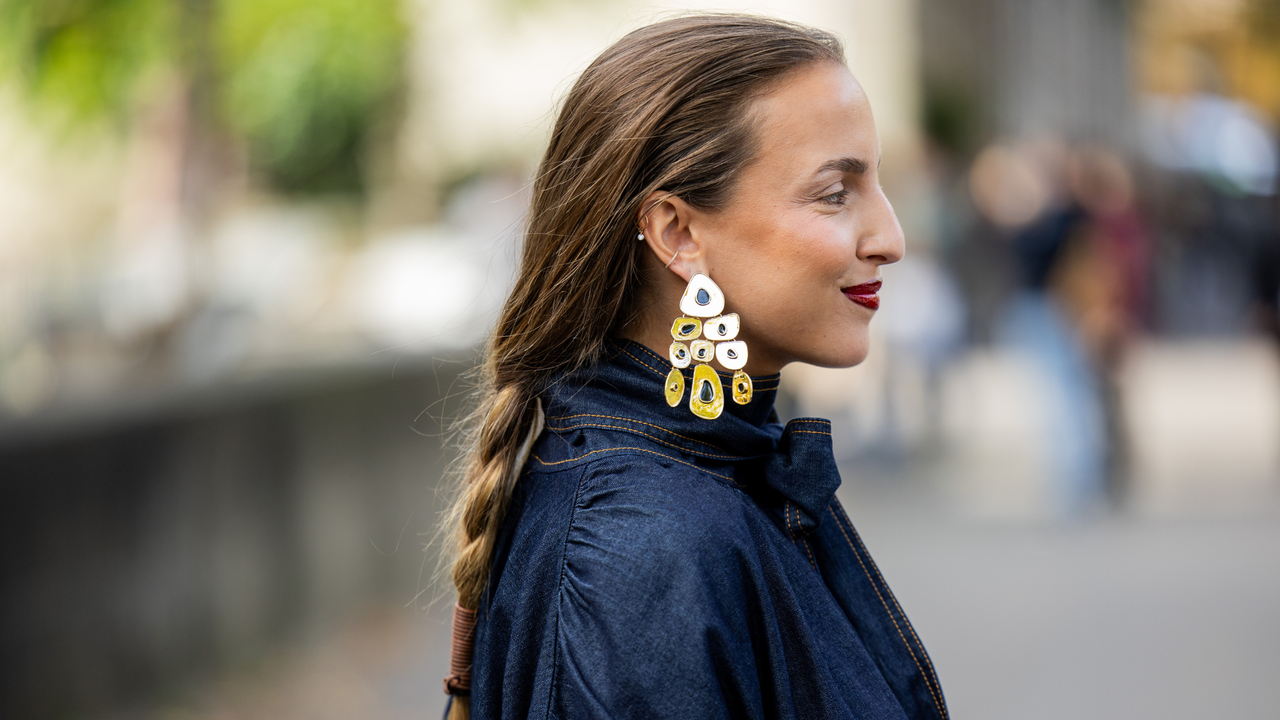 A side on view of a guest at paris fashion week wearing statement earrings, a red lip and a low plait