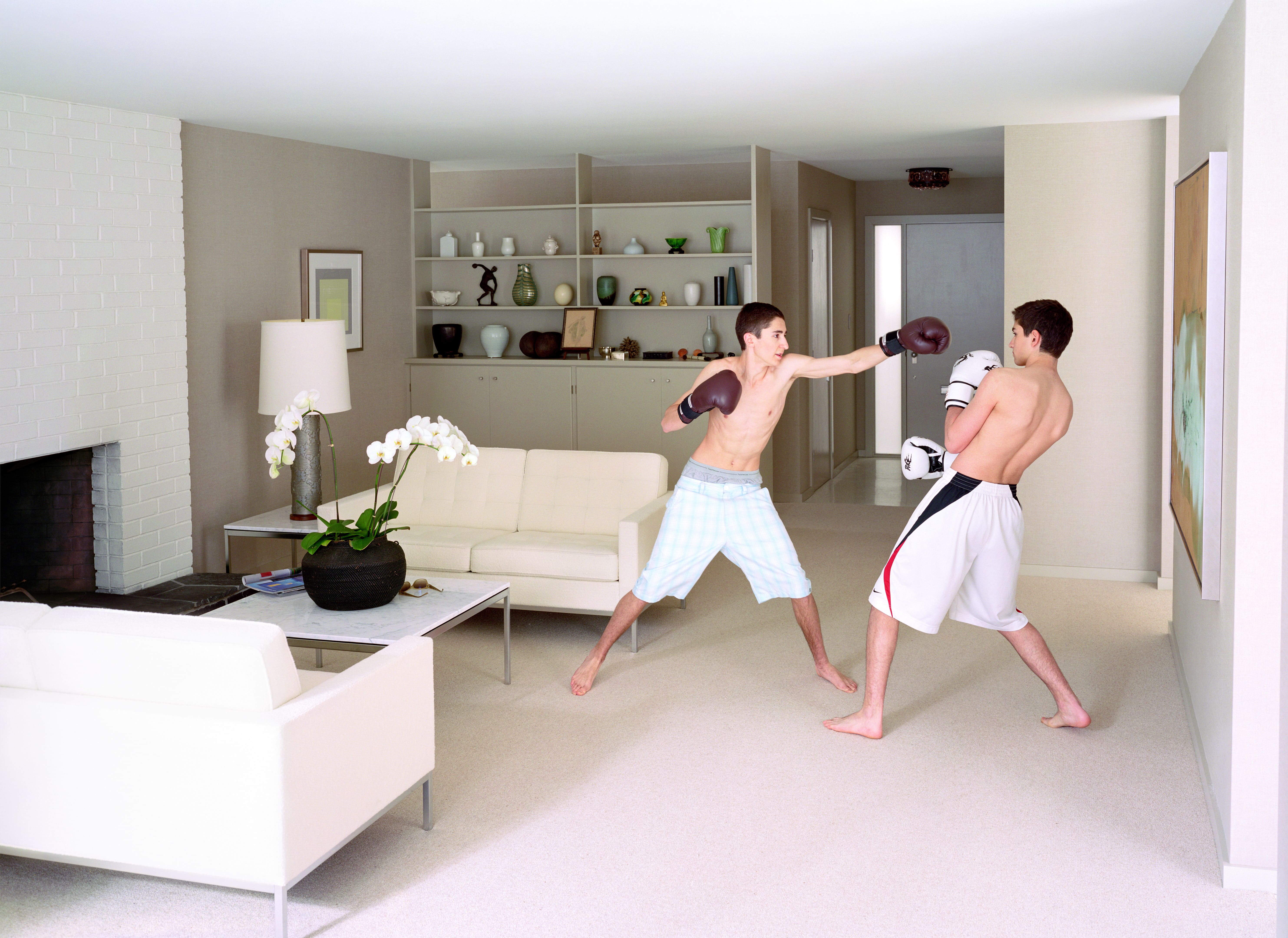 Two young men wearing boxing gloves spar barefoot in a modern, uncluttered living room with white couches and built-in shelving.