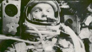 John Glenn, astronaut, is seen from the chest up, sitting in his spacesuit. Two tubes come from out of frame to connect to his helmet. His eyes look left, as the machinery on both sides of his head fills the rest of the photo.