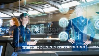 A scene from Star Trek: Discovery. From left to right, we see Sylvia Tilly (young woman with curly, red hair), Jett Reno (older woman with short, gray hair), and Paul Stamets (adult man, with short, light hair). They are all standing in front of a virtual screen checking complicated-looking data.