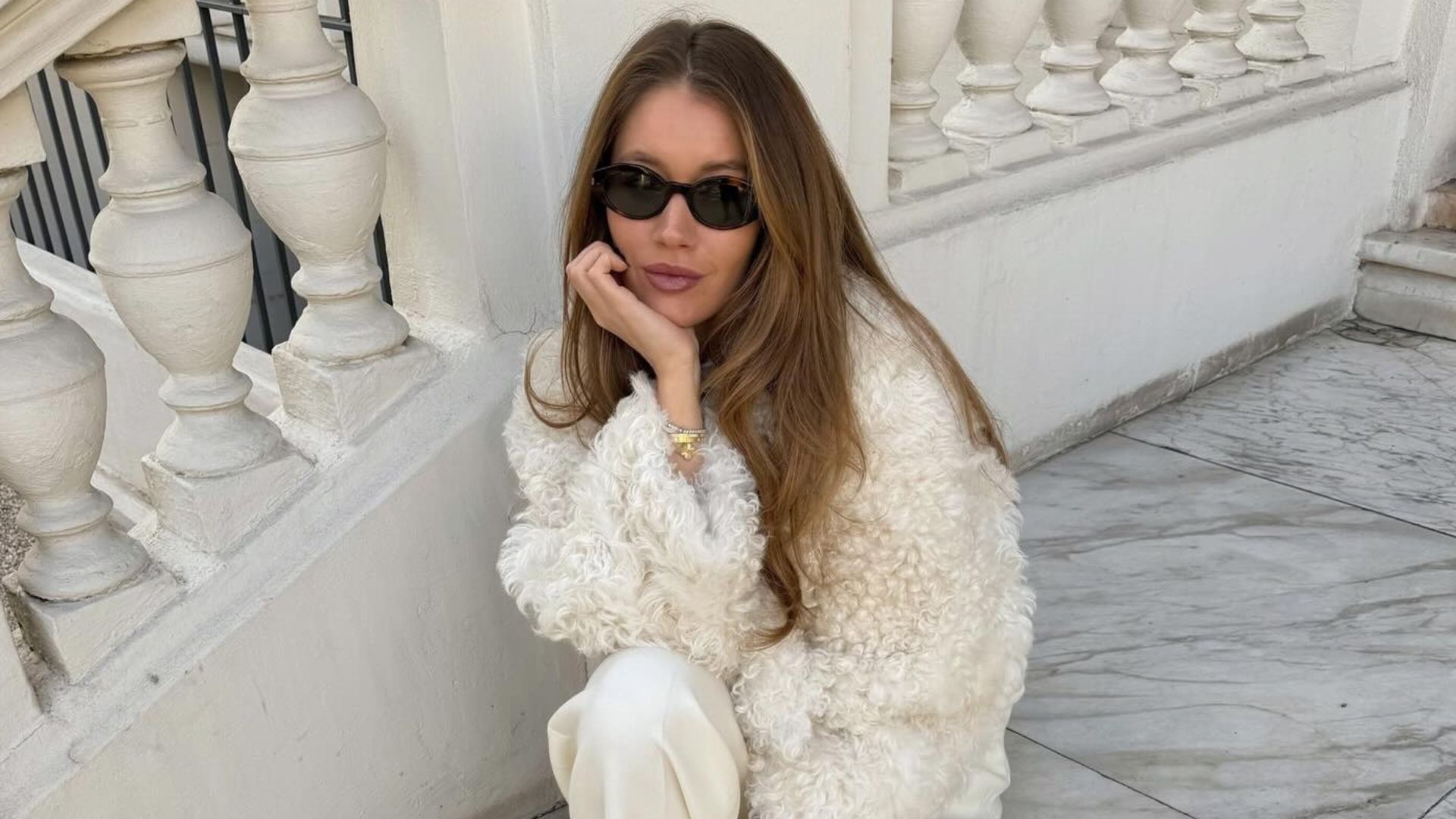 Influencer @lovisabarkman sits on steps outside wearing a cream fur jacket with cream trousers and shearling trainers.