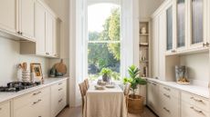 A kitchen in off white tones, with a small dining table placed in the center, with a beige tablecloth on it 