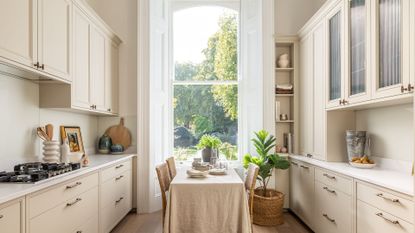 A kitchen in off white tones, with a small dining table placed in the center, with a beige tablecloth on it 