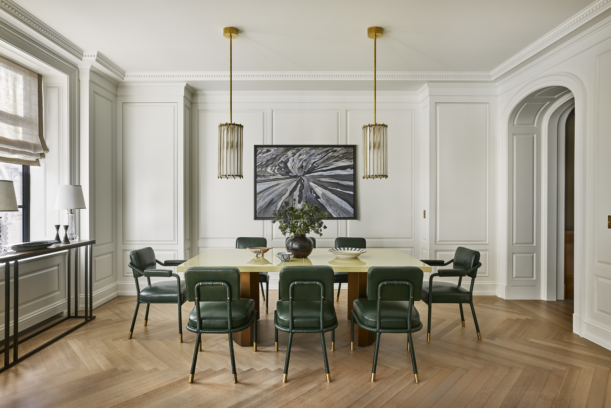 formal dining room with parquetry floors, large table, green leather chairs, two pendant lights, a large artwork, wall molding and an archway through to the next room, a console table under the window dressed with sheer linen roman blinds