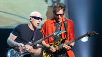 Joe Satriani and Steve Vai, both mid-solo, both playing their signature Ibanez guitars.