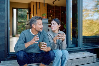 Couple talking and enjoying coffee together on steps outside modern home, feeling happy and connected during leisure time