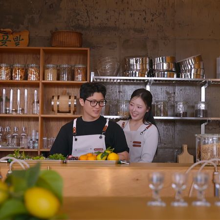A Korean man and woman wear aprons while standing behind a bar in a restaurant kitchen, in Netflix's Korean reality show 'Better Late Than Single' season 2.