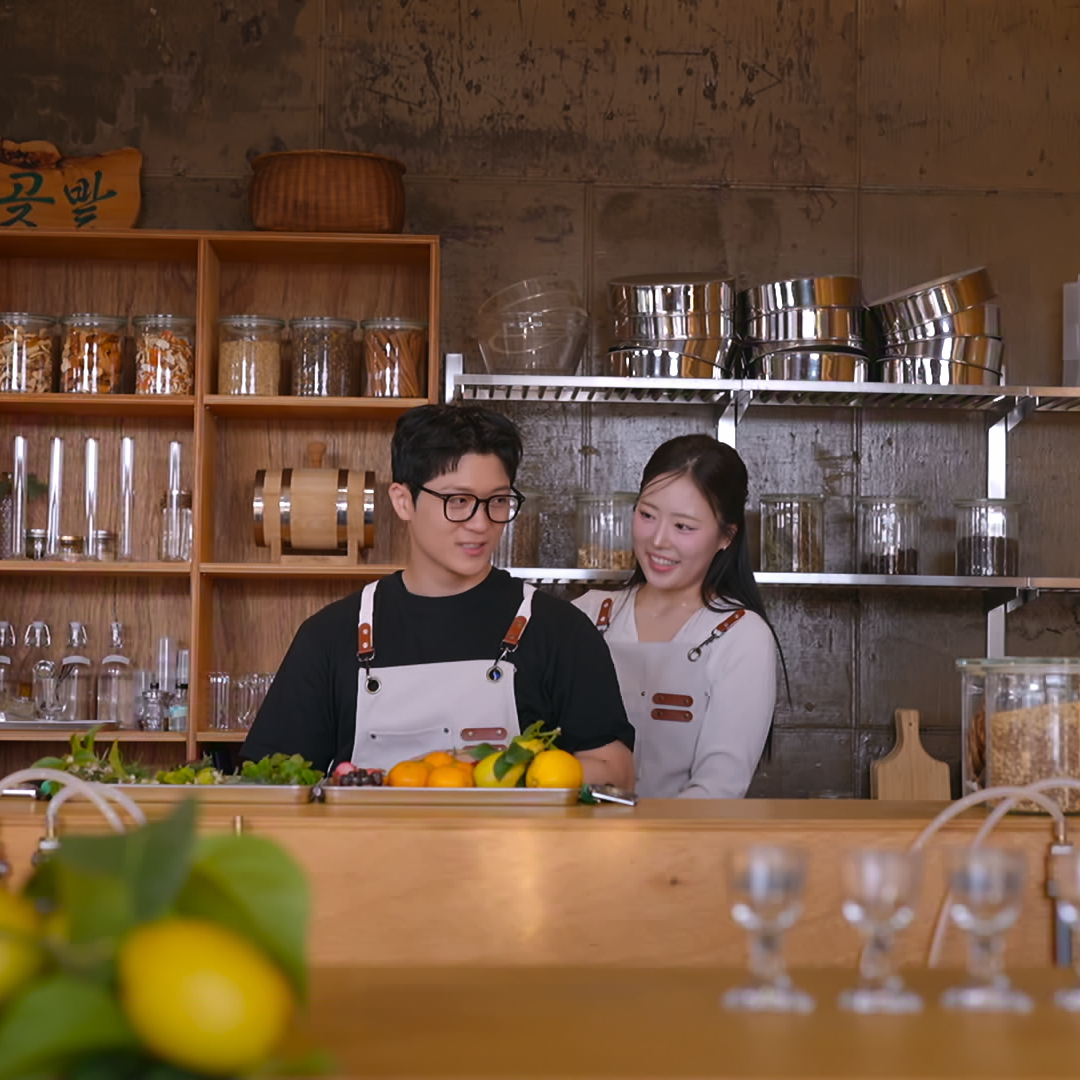 A Korean man and woman wear aprons while standing behind a bar in a restaurant kitchen, in Netflix's Korean reality show 'Better Late Than Single' season 2.