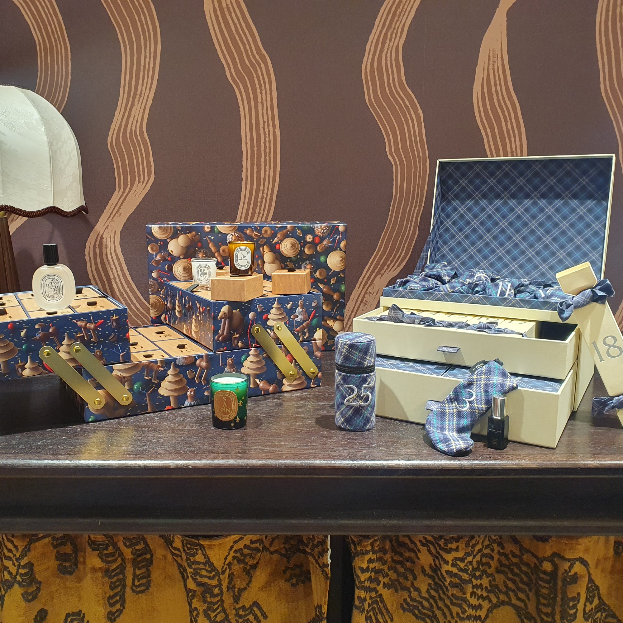 An open Diptyque and Jo Malone luxury advent calendar for 2026 with a few products taken out set on a dark wood table with a brown patterned wallpaper in the background