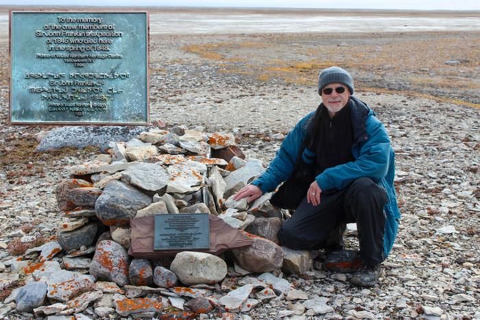 A man in a winter coat and hat kneels on the rocky ground near a tomb covered by rocks and a plaque.
