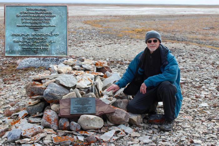 A man in a winter coat and hat kneels on the rocky ground near a  tomb covered by rocks and a plaque. 