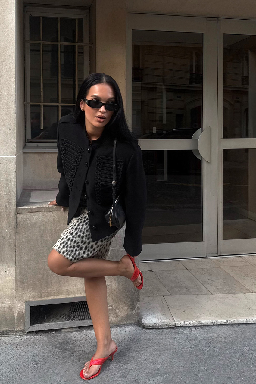 A woman standing outside wearing a black band jacket with a leopard print pencil skirt, red thong sandals, and a black shoulder bag.