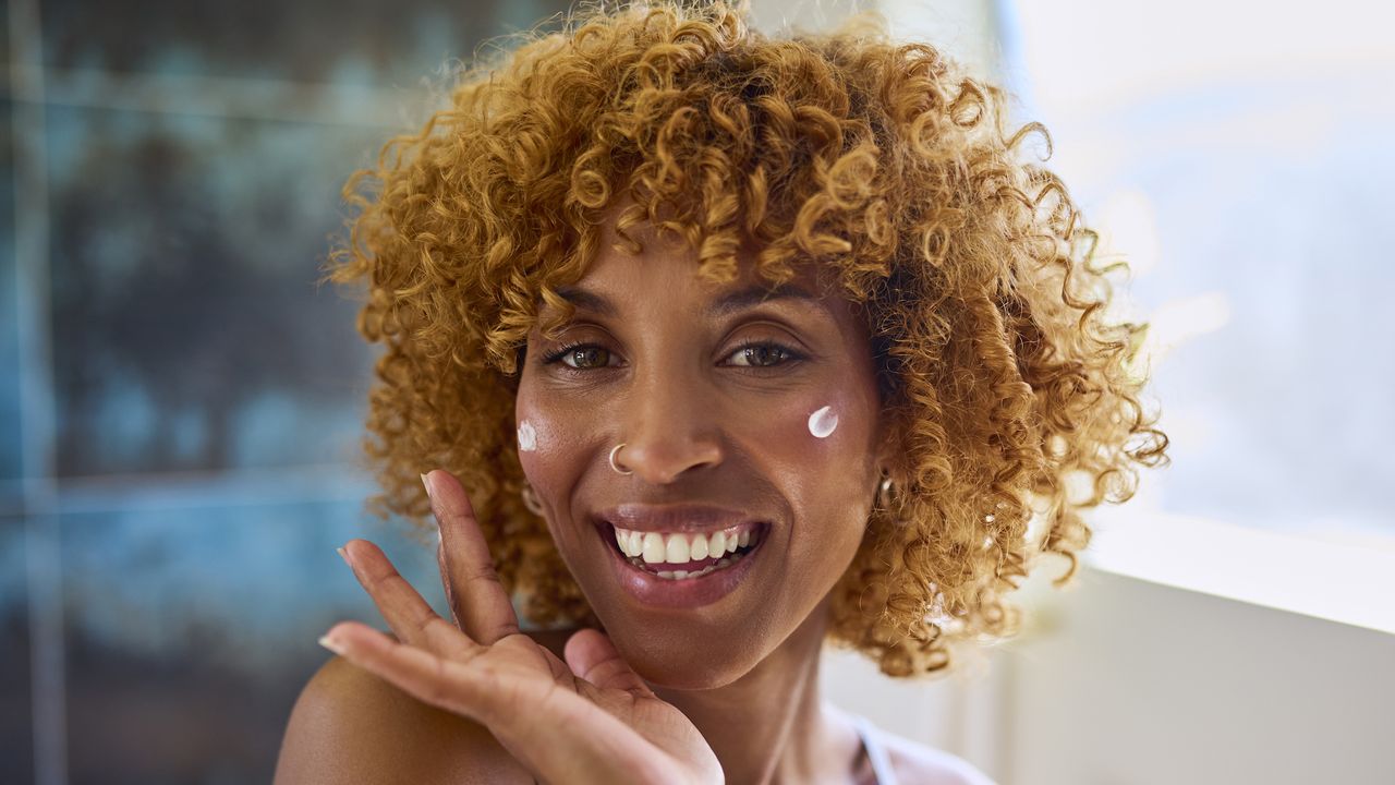 Best moisturiser for mature skin - woman with curly hair applying face cream while smiling