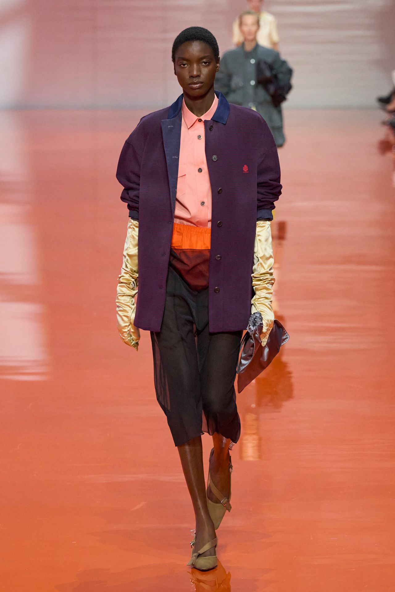 Layering staples for spring 2026 is shown in a photo from Prada's runway show, with a model walking while wearing a purple jacket on top of a salmon button-down shirt that's tucked into orange briefs that a visible through a sheer black skirt styled with a pointed pump, satin gloves, and a satin clutch bag
