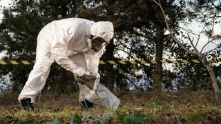 Forensic scientist in a white plastic suit collects a pair of shoes in a plastic bag in a taped off crime scene in the woods