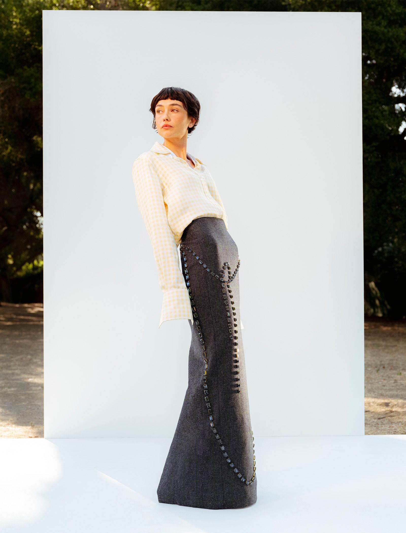 Actress Sydney Chandler stands on a white platform in front of a white wall wearing a Thom Browne yellow and white gingham button-down shirt tucked into a maxi skirt with gem embellishments.
