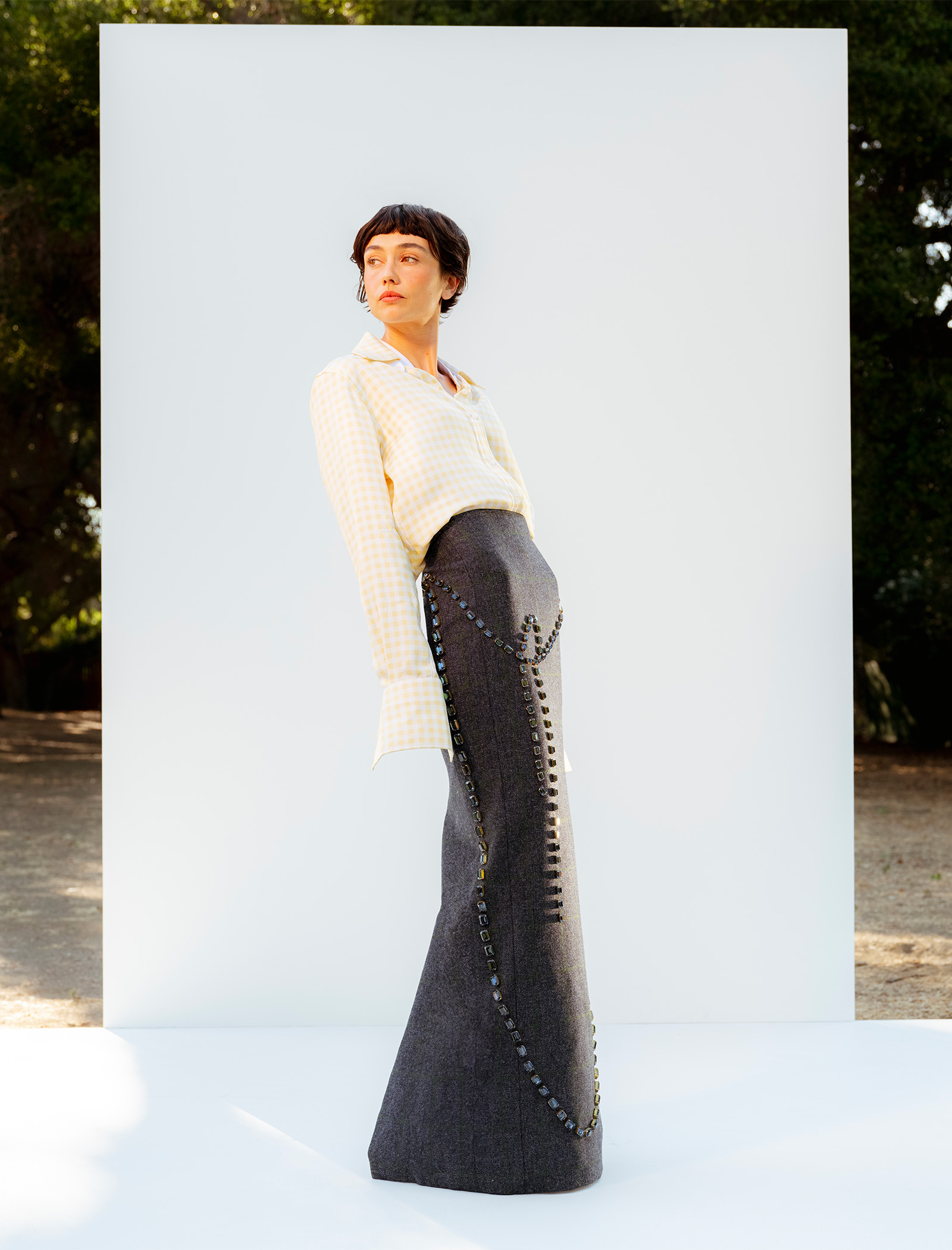 Actress Sydney Chandler stands on a white platform in front of a white wall wearing a Thom Browne yellow and white gingham button-down shirt tucked into a maxi skirt with gem embellishments.