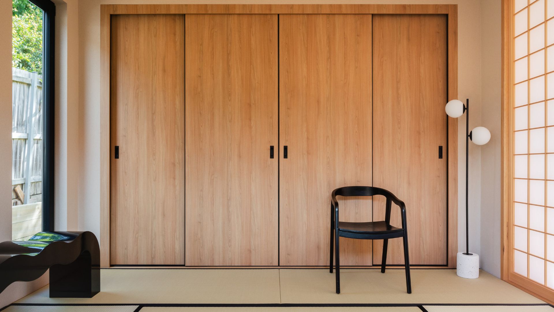 A bright living area with wood sliding doors, a black chair, a black wavy bench, a standing floor lamp, and a set of mats with black trim