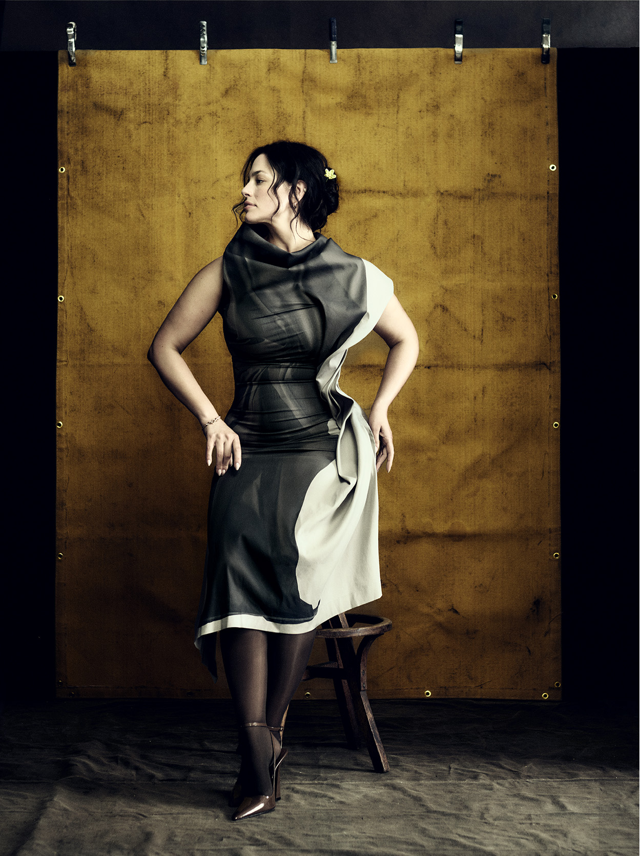 Ashley Graham poses wearing a brown and white couture gown tights and heels sitting on a stool against a yellow backdrop