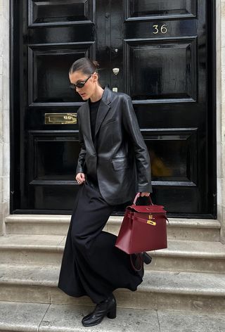 A woman wearing a black leather blazer over a black t-shirt with a black maxi skirt, black ankle boots, and a red handbag.