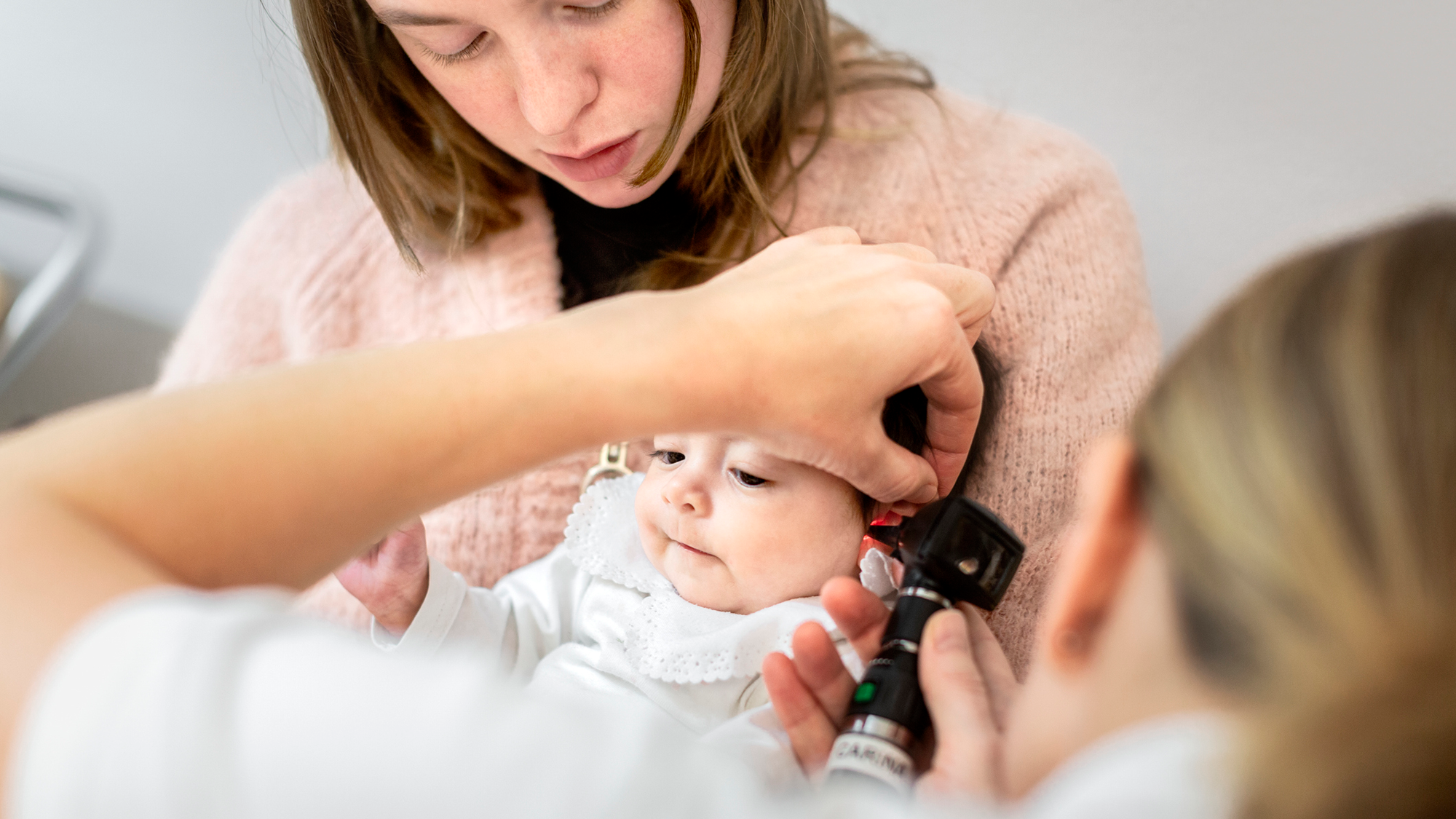 A dark haired woman in a pink sweater holds her infant in front of a woman with a stethoscope